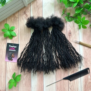 Puede incluir: Extensiones de rastas negras dispuestas sobre una superficie de madera, con un peine negro y una pequeña herramienta. Una tarjeta de visita con "Premium Loc Designs" y una ilustración de rastas también está presente. Hojas verdes y una estructura blanca en el fondo.