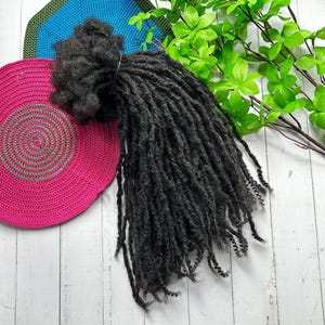 Puede incluir: Un paquete de rastas de pelo humano negro, con algunas hebras sueltas, sobre un fondo de madera blanco.