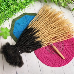 Könnte beinhalten: Ein Set aus zweifarbigen synthetischen Dreadlocks in Schwarz und Blond. Die Dreadlocks werden auf einer rosa gehäkelten Oberfläche mit einem Häkelhaken aus Holz gezeigt.