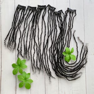 May include: A set of eight black dreadlock hair extensions with clips. The extensions are arranged in a semi-circle on a white wooden surface. There are two green leaves in the lower part of the image.