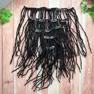 May include: Black dreadlock hair extensions with silver clips, arranged on a wooden surface. The extensions are made of a textured material and are designed to be clipped into natural hair. The dreadlocks vary in length and thickness.