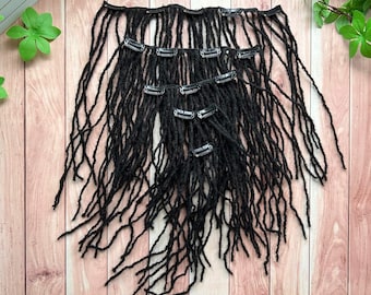 May include: Black dreadlock hair extensions with silver clips, arranged on a wooden surface. The extensions are made of a textured material and are designed to be clipped into natural hair. The dreadlocks vary in length and thickness.