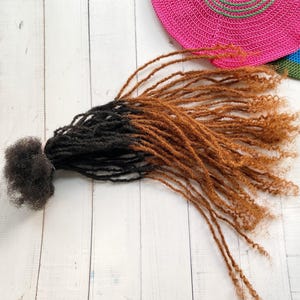 May include: A bundle of brown and black ombre synthetic dreadlocks. The dreadlocks are long and straight with a slight curl at the ends.