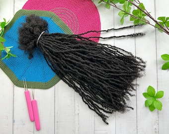 May include: Black synthetic dreadlocks hair extensions on a blue and green crocheted mat. Two pink crochet hooks are on the white wooden surface.