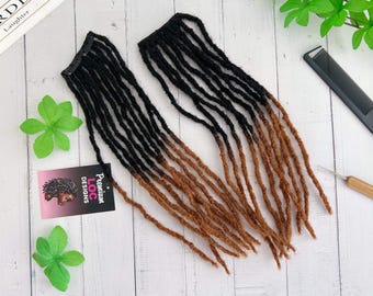 May include: Two bundles of black and brown ombre dreadlock extensions with clips. The dreadlocks are arranged on a white wooden surface, along with a comb, a crochet hook, and a tag that reads "Premium Loc Designs."