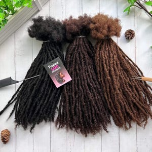May include: Three bundles of synthetic dreadlocks in black, dark brown, and light brown. The dreadlocks are arranged in a fan shape on a white wooden surface. A small tag with the text "Premium Loc Designs" is visible in the image.