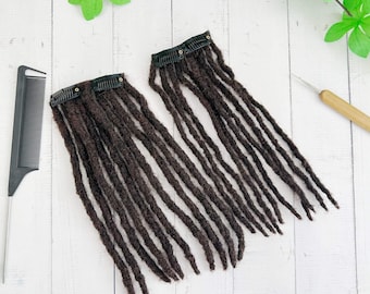 May include: Two sets of dark brown dreadlock extensions with black clips, a black comb, and a crochet hook. The dreadlocks are arranged on a white wooden surface, with green leaves in the background. The dreadlocks are made of a textured material.