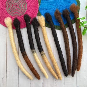 Puede incluir: Una colección de diez rastas sintéticas de varios colores, incluyendo negro, marrón, rubio y marrón claro. Las rastas están dispuestas en una fila sobre una superficie de madera blanca.