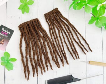 May include: Two bundles of brown dreadlocks are displayed on a white wooden surface, alongside a black comb, a crochet hook, and a business card that reads "Premium Loc Designs". Green leaves and floral accents add a decorative touch.