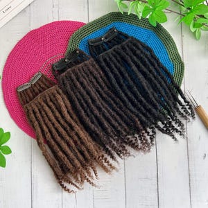 Op de afbeelding: Drie sets dreadlock-extensions in bruin- en zwarttinten, elk met een clip voor eenvoudige bevestiging. De extensions zijn gerangschikt op een wit houten oppervlak, met een roze, blauwe en groene gehaakte cirkel op de achtergrond.