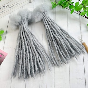 Könnte beinhalten: Zwei Bündel grauer Dreadlocks, jedes oben mit einem flauschigen, strukturierten Material gebunden. Die Dreadlocks sind lang und zylindrisch, mit einem leicht rauen Aussehen. Der Hintergrund ist eine weiße Holzoberfläche.