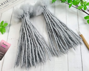 May include: Two bundles of gray dreadlocks, each tied at the top with a fluffy, textured material. The dreadlocks are long and cylindrical, with a slightly rough appearance. The background is a white wooden surface.