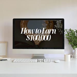 May include: A white computer screen with the text "How to Earn $100,000" displayed on it. The computer is on a white desk with a keyboard, mouse, and a white coffee mug.
