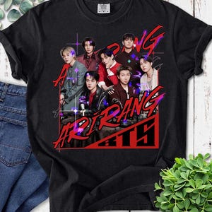 May include: Black t-shirt featuring a graphic of seven people with the text "Art Bang BTS" in red and purple. The design includes a grid pattern and starbursts. The shirt is made of a soft material.