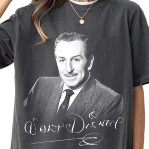 May include: Dark gray t-shirt featuring a black and white portrait of Walt Disney. The shirt has the name "Walt Disney" in a cursive font. The shirt is a vintage style.