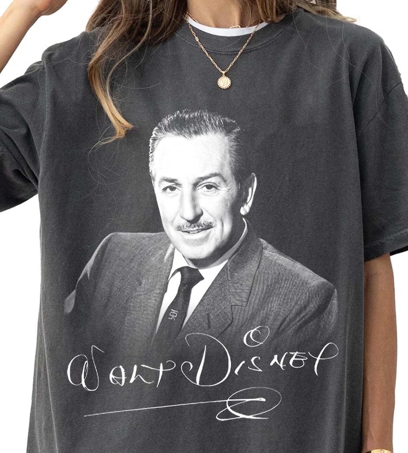 May include: Dark gray t-shirt featuring a black and white portrait of Walt Disney. The shirt has the name "Walt Disney" in a cursive font. The shirt is a vintage style.