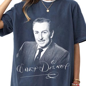 May include: Navy blue t-shirt featuring a black and white portrait of Walt Disney. The shirt has the name "Walt Disney" in a cursive font below the portrait. The shirt is a vintage style.