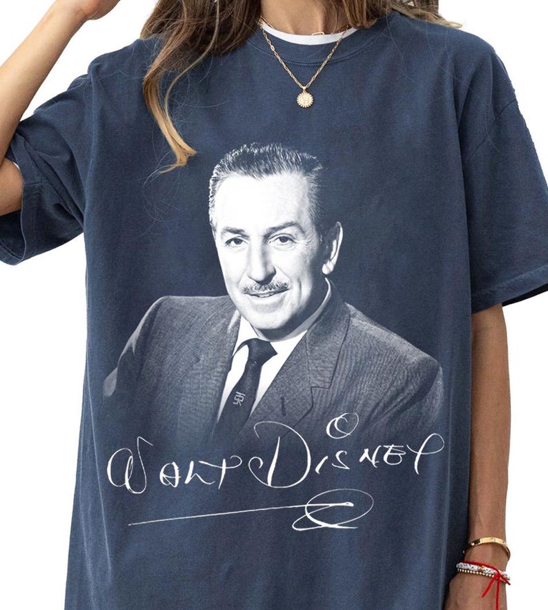 May include: Navy blue t-shirt featuring a black and white portrait of Walt Disney. The shirt has the name "Walt Disney" in a cursive font below the portrait. The shirt is a vintage style.