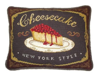 May include: A rectangular decorative pillow with a dark gray background and a stitched image of a slice of cheesecake on a plate. The text "cheesecake" and "New York Style" are embroidered on the pillow. The border is brown and gold.