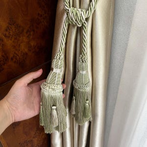 May include: A sage green curtain tieback featuring two decorative tassels with intricate braided rope detailing and a central knot. The tieback is constructed from twisted cord and fine fringe elements, designed to hold a shimmering champagne-colored curtain.