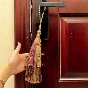 May include: A decorative tassel with a woven top and a long fringe. The tassel features a combination of gold, pink, and burgundy threads. It is hanging on a dark wooden door with a black handle.