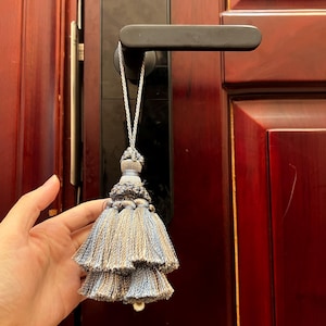 May include: A decorative tassel with a layered design in shades of blue and tan, suspended from a silver cord. The tassel is hanging from a black door handle against a dark red wooden door.