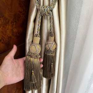 May include: A decorative curtain tieback featuring two long, bronze-colored tassels with intricate braided rope detailing. The tieback is constructed from twisted metallic gold, silver, and bronze cords that form a central knot, connecting to two weighted, cylindrical tassel heads with fine, flowing fringe.