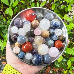 May include: A collection of polished gemstone spheres in a silver bowl. The spheres vary in color, including red, purple, white, blue, and brown. The bowl is held in a hand with a yellow watch band. Green foliage is in the background.