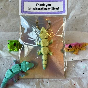 Könnte beinhalten: Ein grüner, blauer und rosa artikulierter Gecko-Schlüsselanhänger in einer durchsichtigen Plastiktüte mit einem Dankeschön-Etikett, auf dem steht: "Vielen Dank, dass Sie mit uns gefeiert haben!"