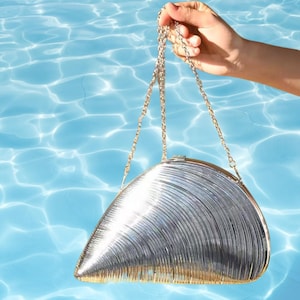 May include: A silver clutch purse with a chain strap. The purse is shaped like a seashell and has a metallic finish.