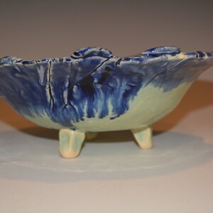 May include: A blue and white ceramic bowl with a wavy rim and three legs. The bowl has a light blue glaze with white streaks and a light green glaze on the bottom.