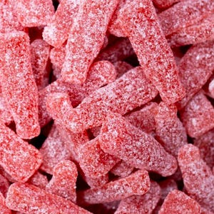 May include: A close-up of a pile of red, sugar-coated candy shaped like bottles. The candy is piled high and the texture is visible.
