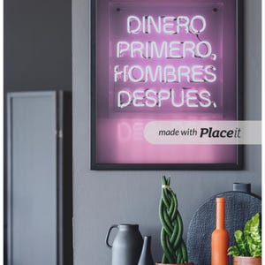 Puede incluir: Un cartel de neón enmarcado con el texto en español "Dinero Primero, Hombres Despues." El cartel es rosa y el texto es blanco. El cartel está colgado en una pared gris con una mesa de madera debajo. Hay tres plantas y un jarrón en la mesa.