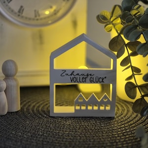 Könnte beinhalten: Ein holzförmiges Hauslicht mit dem Text "Zuhause voller Glück" und einer Reihe von Hausausschnitten. Das Licht befindet sich neben Holzpuppen und einer Uhr. Eine Pflanze befindet sich im Hintergrund.