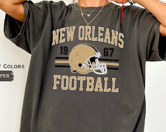 May include: A charcoal grey short-sleeve t-shirt featuring the text NEW ORLEANS FOOTBALL in beige, with the numbers 19 and 67 flanking a beige football helmet graphic. The shirt includes the printed text COMFORT COLORS PEPPER on the side.