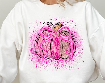Op de afbeelding: Een wit sweatshirt met lange mouwen en een centrale afbeelding van een pompoen versierd met roze luipaardprint, glittertexturen en linten voor borstkankerbewustzijn, vastgebonden met een grote roze strik op een achtergrond van roze verfspatten.