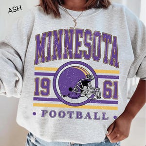 May include: An ash gray crewneck sweatshirt featuring a distressed purple and yellow graphic. The text reads MINNESOTA 1961 FOOTBALL, centered around a purple football helmet illustration. The design has a vintage, weathered texture throughout.