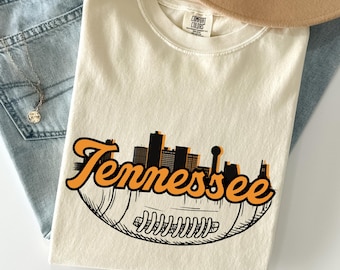 Könnte beinhalten: Ein cremefarbenes Kurzarm-T-Shirt mit einer Grafik eines Footballs, über dem sich die schwarze Silhouette einer Skyline befindet. Das Wort Tennessee ist in großen, orangefarbenen Schreibschriftbuchstaben über die Mitte der Grafik gedruckt.