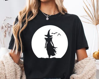 Puede incluir: Una camiseta negra con un gran gráfico circular blanco en el pecho. El gráfico muestra la silueta de una bruja con sombrero puntiagudo y vestido largo, de pie frente a una luna llena con dos pequeños murciélagos volando a la derecha.