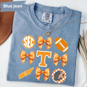 A blue t-shirt with various orange and white graphics and text, including a large "T" with a bow, a football, and a dog.