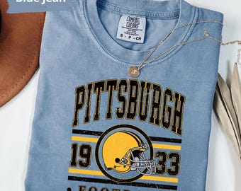 May include: A blue jean coloured short-sleeve t-shirt with a crew neckline. The graphic features the text PITTSBURGH in black with yellow outlines, a yellow football helmet, the number 1933, and the word FOOTBALL in black lettering.