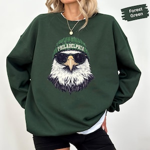 May include: A forest green crewneck sweatshirt featuring a graphic of a bald eagle wearing a green knit beanie with the text PHILADELPHIA and black sunglasses. The eagle has a white head, yellow beak, and dark feathers around the neck.