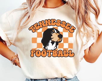 May include: A cream-colored t-shirt featuring a graphic of a black and white dog head centered over an orange and white checkered pattern. The text "TENNESSEE" is arched above the dog, and "FOOTBALL" is printed below in bold orange lettering.