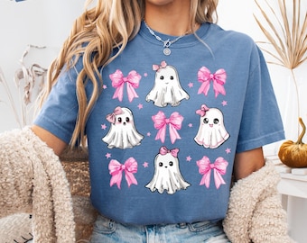 May include: A blue jean coloured t-shirt featuring a graphic print of four white ghosts and five pink bows arranged in a grid pattern, accented with small pink stars. The text "BLUE JEAN" appears in a small box in the upper left corner.