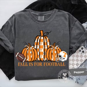 May include: A charcoal gray crewneck t-shirt featuring a graphic of three orange pumpkins, one with a white checkerboard pattern and a T logo. A brown football and a white football helmet are included, with the text FALL IS FOR FOOTBALL below.