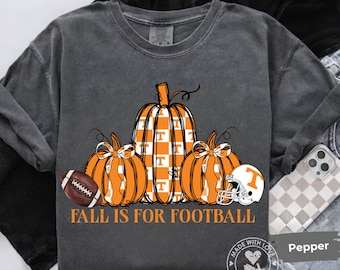 May include: A charcoal gray crewneck t-shirt featuring a graphic of three orange pumpkins, one with a white checkerboard pattern and a T logo. A brown football and a white football helmet are included, with the text FALL IS FOR FOOTBALL below.