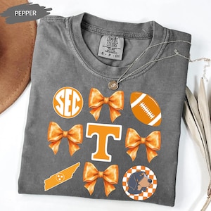 May include: A charcoal grey t-shirt featuring a graphic print with orange elements including the SEC logo, four orange bows, a football, a Tennessee state outline, a large letter T, and a dog head inside an orange and white checkered circle.