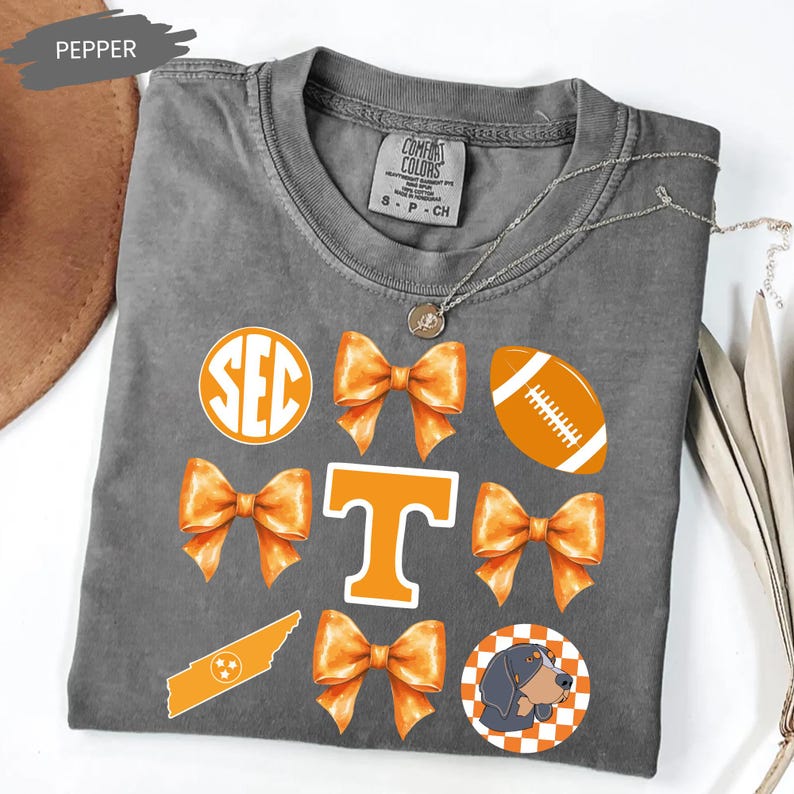 A gray t-shirt with various orange and white graphics and text, including a large "T" with a bow, a football, and a dog.