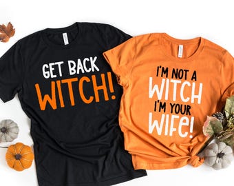 Puede incluir: Una camiseta negra con el texto naranja GET BACK WITCH! y una camiseta naranja con el texto blanco I'M NOT A WITCH I'M YOUR WIFE!. Ambas camisetas tienen mangas cortas y cuello redondo, presentadas junto a pequeñas calabazas decorativas.