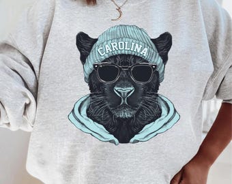 May include: An ash grey crewneck sweatshirt featuring a graphic illustration of a black panther wearing a light blue beanie with the text CAROLINA printed on it, black sunglasses, and a light blue hooded sweatshirt layered underneath.